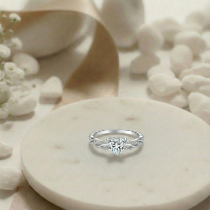 Silver ring with a heart-shaped center stone on a marble surface with flowers.