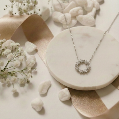 Silver necklace with a circular pendant on a marble surface with small white flowers and stones.