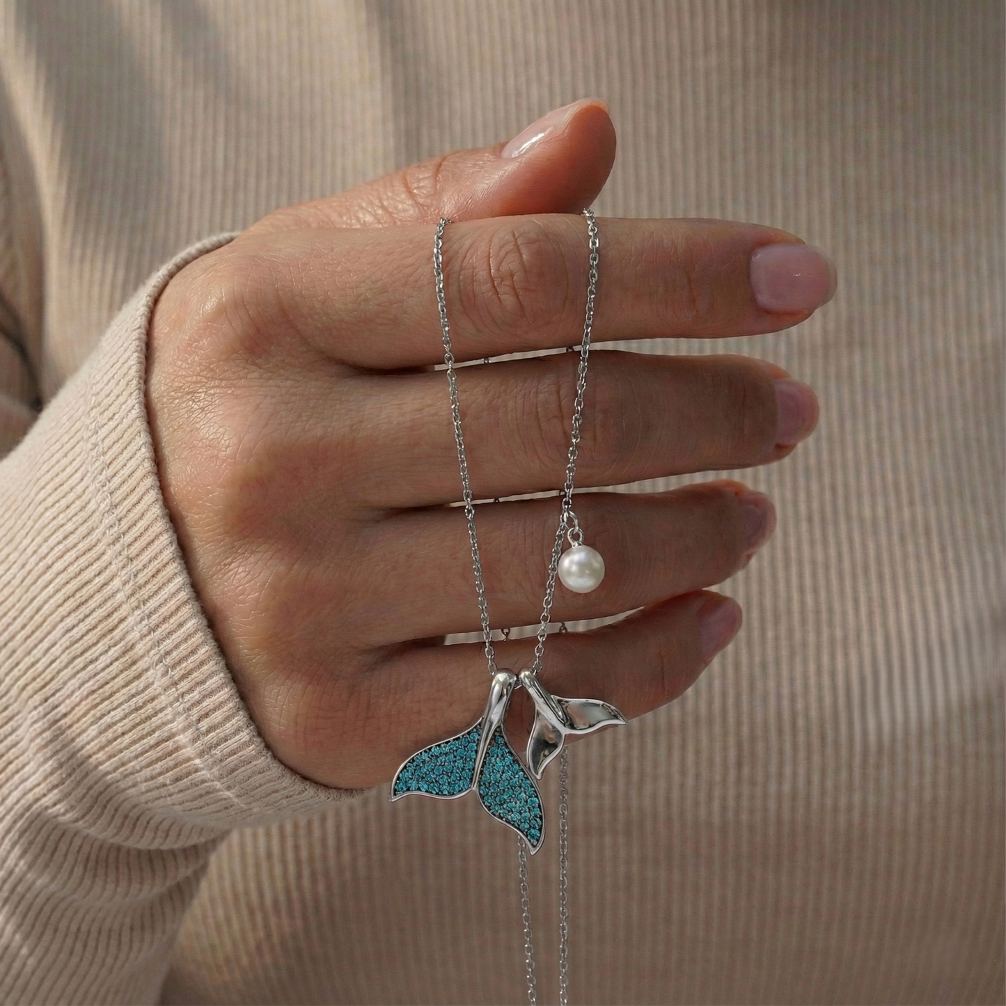 Hand holding a necklace with a blue pendant and pearl, against a beige background