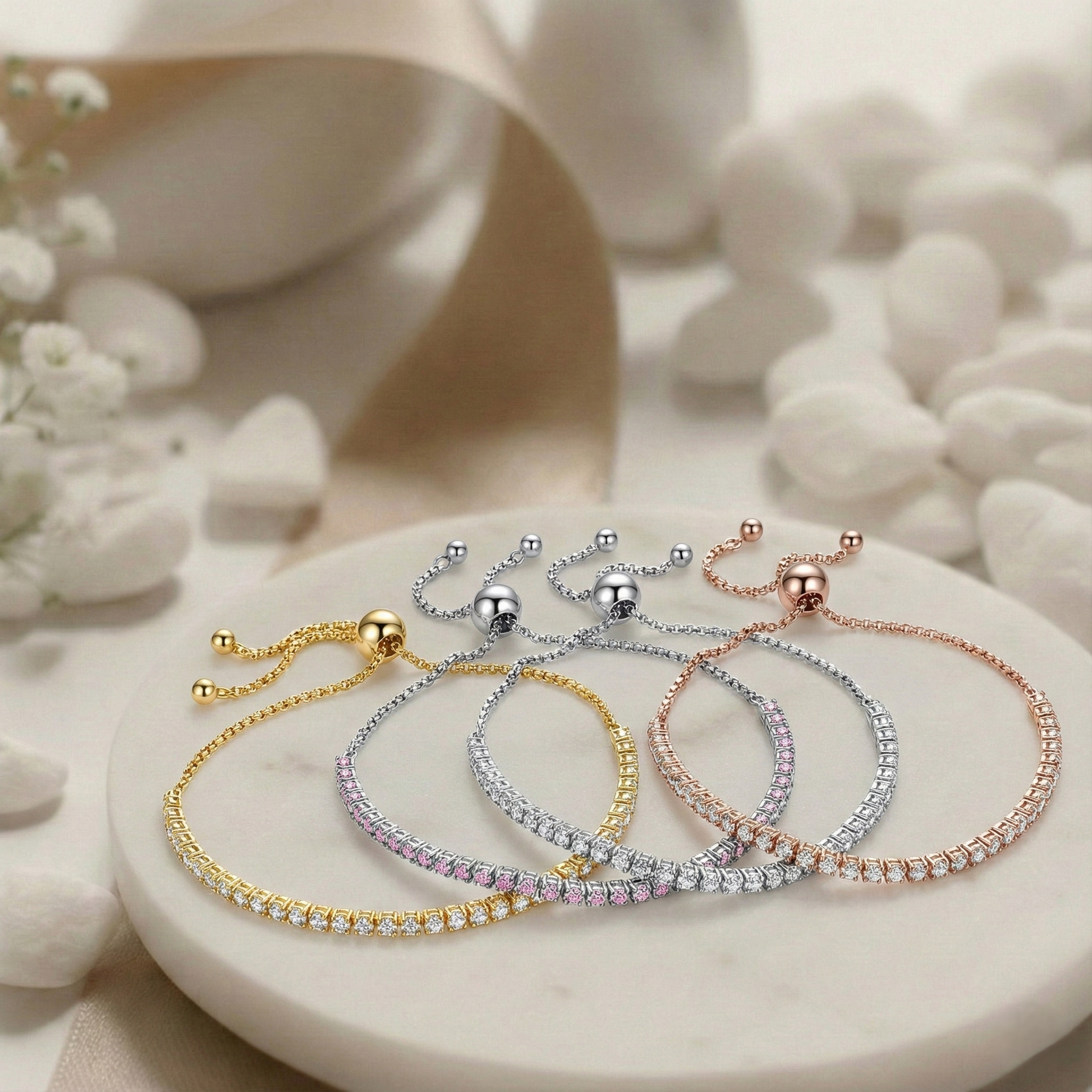Four bracelets in gold, silver, and rose gold on a white stone surface with flowers.