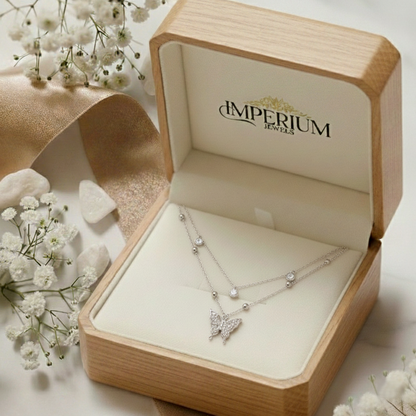 Butterfly necklace in a wooden jewelry box with 'Imperium Jewels' branding, surrounded by white flowers.