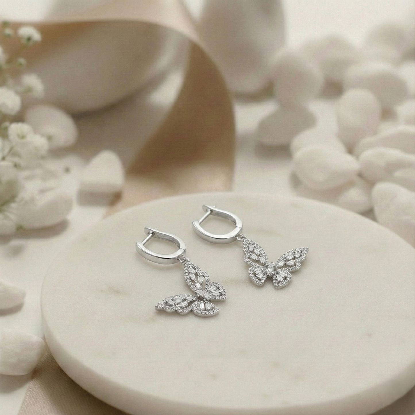 Silver earrings with leaf design on a white stone surface with flowers and shells in the background