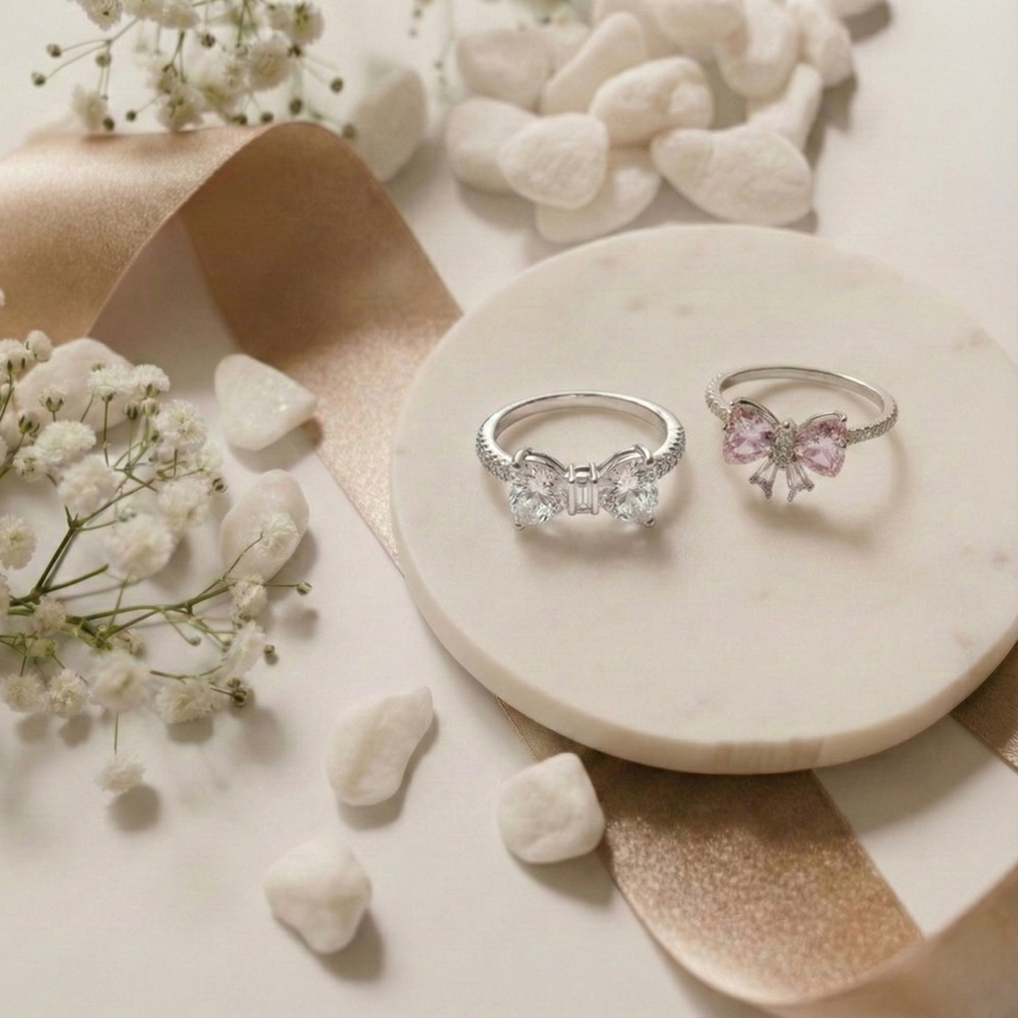 Two silver rings with bow designs on a marble surface with flowers and stones.