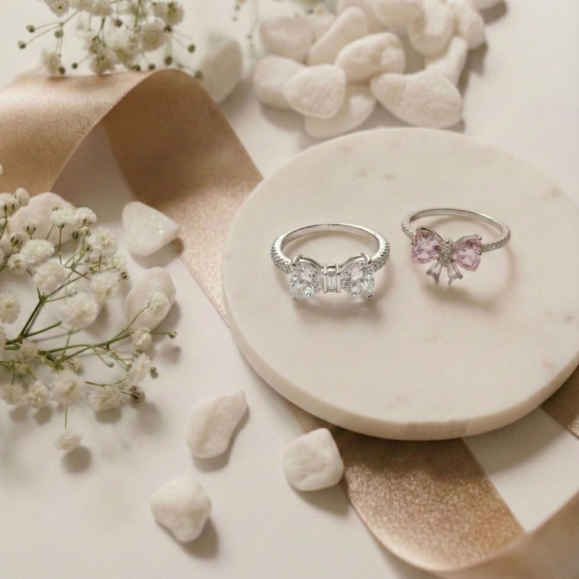 Two silver rings with bow designs on a marble surface with flowers and stones.